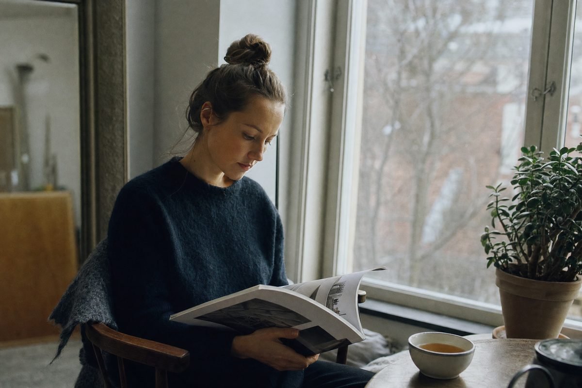 A person reading a printed magazine at a window seat with a cup of tea